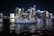 A luxurious yacht sails on a calm body of water against the backdrop of a vibrant city skyline illuminated at night. The high-rise buildings are adorned with bright neon lights, reflecting vivid colors on the water surface.