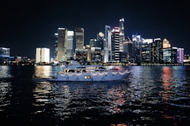A luxurious yacht sails on a calm body of water against the backdrop of a vibrant city skyline illuminated at night. The high-rise buildings are adorned with bright neon lights, reflecting vivid colors on the water surface.
