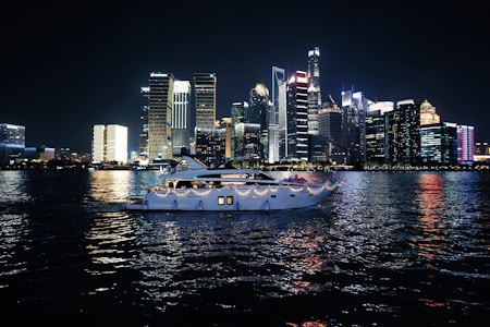 A luxurious yacht sails on a calm body of water against the backdrop of a vibrant city skyline illuminated at night. The high-rise buildings are adorned with bright neon lights, reflecting vivid colors on the water surface.