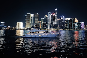 A luxurious yacht sails on a calm body of water against the backdrop of a vibrant city skyline illuminated at night. The high-rise buildings are adorned with bright neon lights, reflecting vivid colors on the water surface.