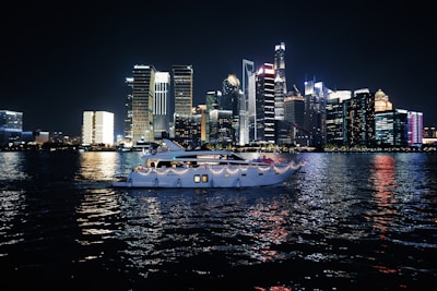 A luxurious yacht sails on a calm body of water against the backdrop of a vibrant city skyline illuminated at night. The high-rise buildings are adorned with bright neon lights, reflecting vivid colors on the water surface.