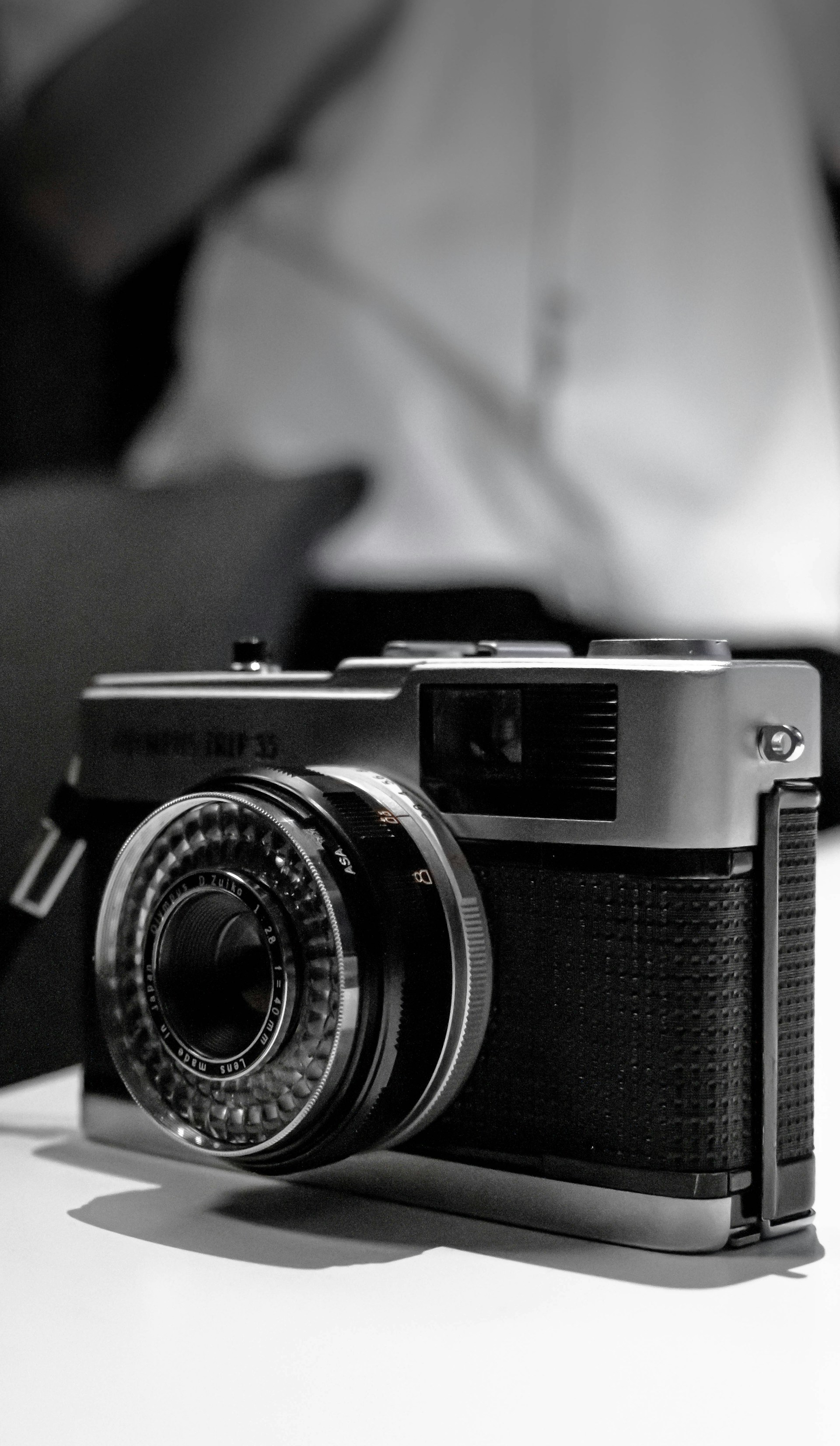 A vintage camera with a classic design and textured grip rests on a surface. Its circular lens and metallic details stand out against a slightly blurred background.