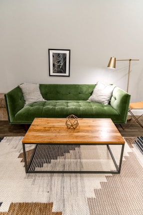 A modern living room features a green velvet sofa with two gray pillows. Above the sofa hangs a framed abstract artwork. In front of the sofa is a wooden coffee table with a geometric sculpture on top. The room includes a tall gold floor lamp, a patterned rug, and an ornate chair.