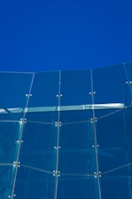 A modern glass facade composed of tall, vertical glass panels connected by metallic fixtures. The glass reflects the clear blue sky, creating a harmonious blend of transparency and solidity.