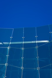 A modern glass facade composed of tall, vertical glass panels connected by metallic fixtures. The glass reflects the clear blue sky, creating a harmonious blend of transparency and solidity.