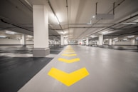 An empty underground parking garage with a clean, polished floor marked with yellow directional arrows. The space is well-lit with fluorescent lights and features concrete columns evenly spaced along the center.