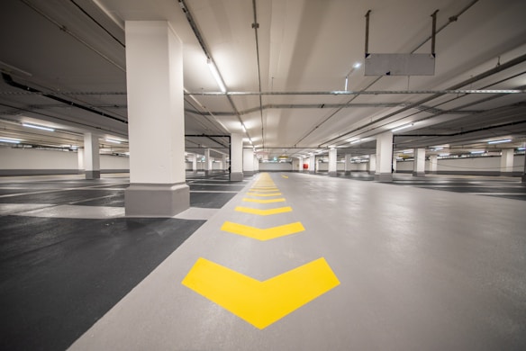 An empty underground parking garage with a clean, polished floor marked with yellow directional arrows. The space is well-lit with fluorescent lights and features concrete columns evenly spaced along the center.