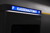 A parking payment machine is partially visible, with a brightly lit sign in blue displaying the word 'Kassenautomat' and the letter 'P' on both sides. The surrounding area is dimly lit, emphasizing the illuminated sign.
