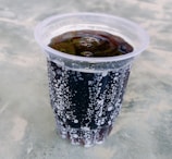 A plastic cup filled with a dark carbonated beverage, displaying a layer of bubbles on the surface and inside the liquid. The background consists of a grayish textured surface, possibly a table.