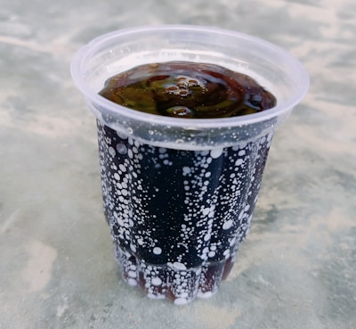 A plastic cup filled with a dark carbonated beverage, displaying a layer of bubbles on the surface and inside the liquid. The background consists of a grayish textured surface, possibly a table.