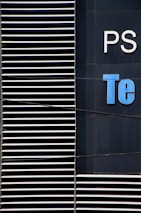 A close-up view of a modern building facade with a series of horizontal white slats on the left and partial letters 'PS' and 'Te' in white and blue on a dark surface on the right.