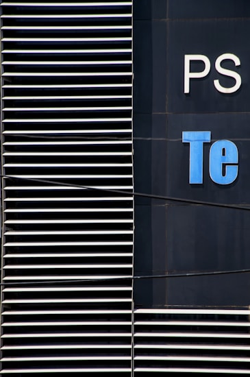 A close-up view of a modern building facade with a series of horizontal white slats on the left and partial letters 'PS' and 'Te' in white and blue on a dark surface on the right.