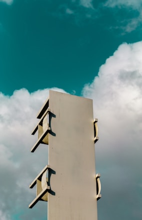 A tall, rectangular metal structure with curved brackets is set against a backdrop of a cloudy sky. The structure appears industrial and possibly serves as a support or part of a larger system. The sky provides a contrast with its mix of dark turquoise and white clouds.