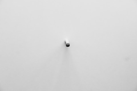A minimalistic scene featuring a white surface with a small, dark, cylindrical object protruding from the center. The shadow created by the object adds depth to the otherwise plain background.