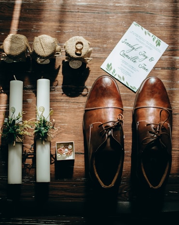 A flat lay arrangement featuring a pair of brown leather shoes, two white candles with green decorative elements, small burlap-wrapped jars with a gold wedding ring on top, a white card with green leaves, and a small box containing jewelry with gemstones and pearls. The items are placed on a wooden surface with sunlight casting shadows.