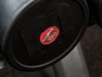 A black weight plate with a red center label displaying the brand name 'BodyMax' and the weight '30kg'. The weight appears attached to a piece of gym equipment.