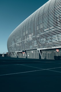 A large, modern architectural structure with a curved facade composed of horizontal slats, possibly part of a stadium or an arena. The structure features a sleek, metallic appearance with sparse signs and infrastructure in the surrounding area, suggesting an urban environment.