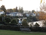 A scenic view of a residential area with houses nestled among lush greenery. A Canadian flag is visible, symbolizing the location, and there is a bench in the foreground on a grassy area. The scene is set against a backdrop of tall trees, with a body of water reflecting the light. The autumn foliage adds a warm tone to the overall atmosphere.