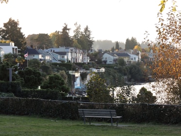A scenic view of a residential area with houses nestled among lush greenery. A Canadian flag is visible, symbolizing the location, and there is a bench in the foreground on a grassy area. The scene is set against a backdrop of tall trees, with a body of water reflecting the light. The autumn foliage adds a warm tone to the overall atmosphere.