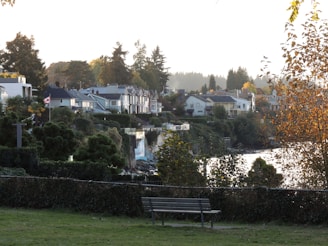 A scenic view of a residential area with houses nestled among lush greenery. A Canadian flag is visible, symbolizing the location, and there is a bench in the foreground on a grassy area. The scene is set against a backdrop of tall trees, with a body of water reflecting the light. The autumn foliage adds a warm tone to the overall atmosphere.