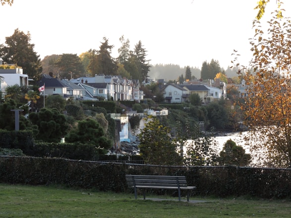 A scenic view of a residential area with houses nestled among lush greenery. A Canadian flag is visible, symbolizing the location, and there is a bench in the foreground on a grassy area. The scene is set against a backdrop of tall trees, with a body of water reflecting the light. The autumn foliage adds a warm tone to the overall atmosphere.