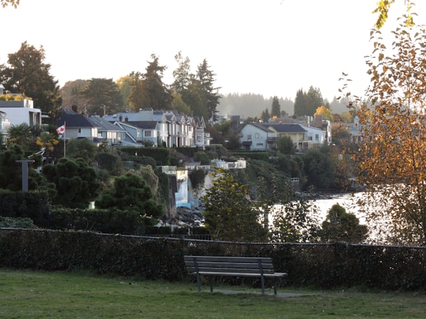 A scenic view of a residential area with houses nestled among lush greenery. A Canadian flag is visible, symbolizing the location, and there is a bench in the foreground on a grassy area. The scene is set against a backdrop of tall trees, with a body of water reflecting the light. The autumn foliage adds a warm tone to the overall atmosphere.