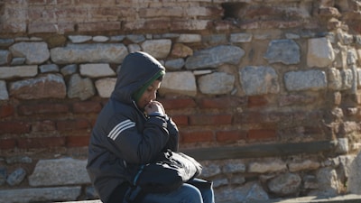 A young child sits alone against a rustic stone wall, wearing a dark hooded jacket. The child appears deep in thought, with their head slightly bowed and hands brought close to their face, as if seeking warmth.