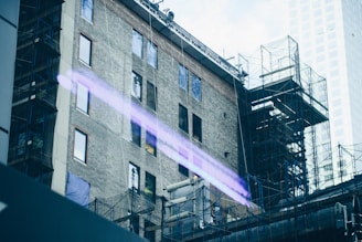 A partially constructed or renovated urban building with visible scaffolding and support structures. The building facade is composed of brick and features several windows. There is a prominent purple light streak running across the image, likely a reflection or digital artifact. The background shows a taller, modern glass building.