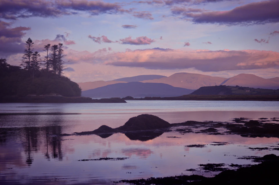 The image depicts a serene lake scene at sunset, with a large rock formation on one side and mountains rising into the distance beyond it. The sky is painted in hues of pink and purple, creating a beautiful backdrop for the tranquil waters below. The perspective from the shoreline offers a panoramic view of the landscape, showcasing the natural beauty of the scene.