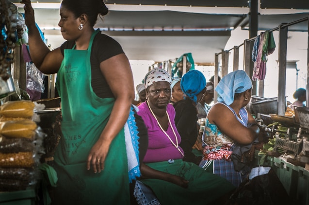 A busy market scene with several women working at stalls lined with bags of goods. The women are wearing colorful headscarves and aprons as they interact with the products and each other. The lighting suggests an indoor environment, possibly a traditional market setting.