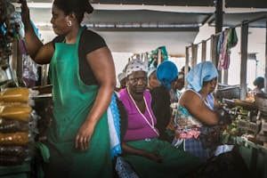 A busy market scene with several women working at stalls lined with bags of goods. The women are wearing colorful headscarves and aprons as they interact with the products and each other. The lighting suggests an indoor environment, possibly a traditional market setting.
