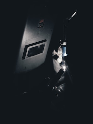 A person is welding in a dark environment. The welding torch creates a bright spark, illuminating part of the welder's protective gear and a welding mask. The scene appears industrial with a strong contrast between light and shadow.