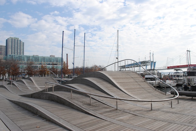 A modern urban landscape featuring a wooden boardwalk with undulating, wave-like designs. Several sailboats are docked nearby, with masts visible against the sky. Building structures, including high-rise apartments and a bridge, are present in the background, framed by sparse trees.