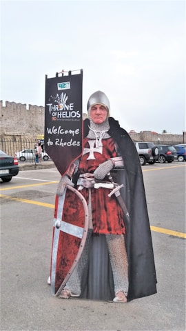 A cutout figure of a medieval knight dressed in chainmail and a red tunic, holding a shield and standing in front of a sign that reads 'Throne of Helios 9D Experience' with 'Welcome to Rhodes' underneath. Behind the cutout, a stone fortress wall and parked cars are visible.