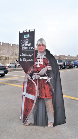A cutout figure of a medieval knight dressed in chainmail and a red tunic, holding a shield and standing in front of a sign that reads 'Throne of Helios 9D Experience' with 'Welcome to Rhodes' underneath. Behind the cutout, a stone fortress wall and parked cars are visible.