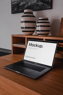 A laptop, possibly a MacBook Pro, is placed on a wooden table in front of two large ceramic vases or jars with black and white stripes. The screen displays a mockup design with text. Behind, there is a framed black and white photograph on the wall.