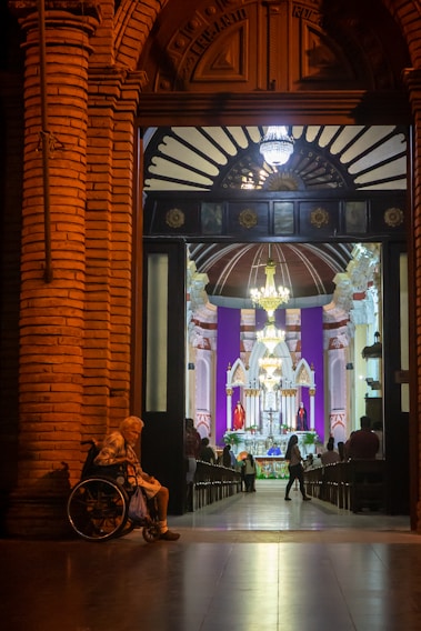 A warmly lit, ornate doorway opens into a church interior, where a congregation gathers near a brightly illuminated altar. An elderly person in a wheelchair sits on the left side of the entrance.