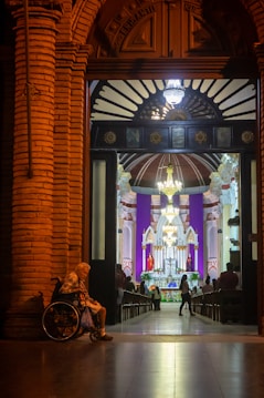 A warmly lit, ornate doorway opens into a church interior, where a congregation gathers near a brightly illuminated altar. An elderly person in a wheelchair sits on the left side of the entrance.