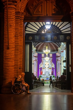 A warmly lit, ornate doorway opens into a church interior, where a congregation gathers near a brightly illuminated altar. An elderly person in a wheelchair sits on the left side of the entrance.