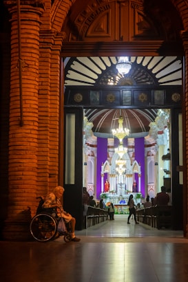 A warmly lit, ornate doorway opens into a church interior, where a congregation gathers near a brightly illuminated altar. An elderly person in a wheelchair sits on the left side of the entrance.