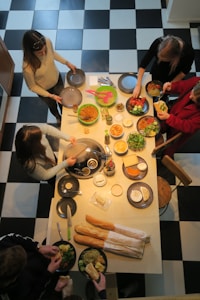 Several people gather around a kitchen island with assorted dishes and ingredients like salad, pasta, bread, cheese, and various vegetables. The countertop is adorned with plates and utensils, and the room features a black and white checkered floor.