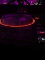 A DJ controller with illuminated buttons in a dark setting featuring a prominently glowing turntable surrounded by a red light. The background is blurred with hints of purple and magenta lights.