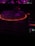 A DJ controller with illuminated buttons in a dark setting featuring a prominently glowing turntable surrounded by a red light. The background is blurred with hints of purple and magenta lights.