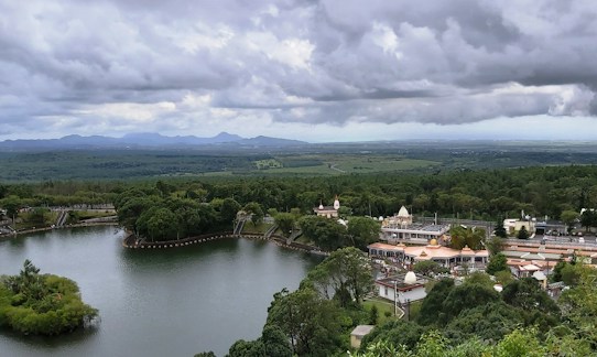 A scenic view features a large body of water surrounded by lush greenery and trees. On the right side, a cluster of buildings and a temple-like structure are nestled among the trees. In the distance, rolling hills and vast plains extend towards the horizon, under a dramatic cloudy sky.
