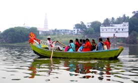 A small green boat is carrying a group of people, predominantly women in colorful traditional clothing, on a calm body of water. The boatman is rowing with a long wooden oar. In the background, there are trees, a large white building, and a tall, slender temple structure. The water reflects the boat and the buildings.