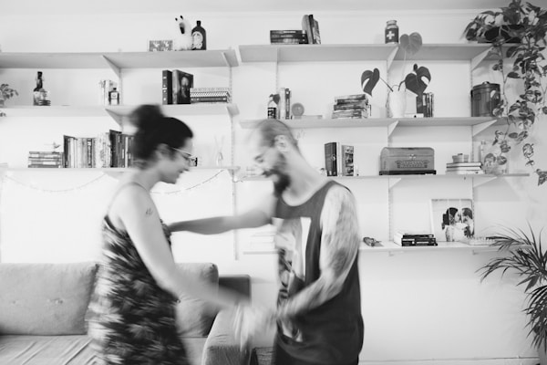 A black and white photograph capturing a blurred moment of two people, possibly dancing or moving energetically, in a cozy living room. The space has several shelves filled with books, plants, and decorative items. A couch and some framed photos are visible, creating a homely atmosphere.