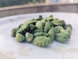 A close-up image of several green cardamom pods placed on a white surface, possibly a plate. The background is blurred, drawing attention to the cardamom pods in the foreground.