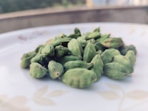 A close-up image of several green cardamom pods placed on a white surface, possibly a plate. The background is blurred, drawing attention to the cardamom pods in the foreground.
