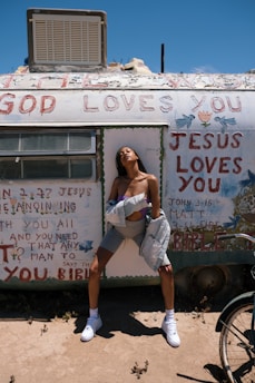 A person stands confidently in front of a weathered trailer with religious messages painted across it. The sky is clear and bright, and the individual is dressed in casual clothing, including sneakers, shorts, and a jacket draped off their shoulders.