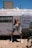 A person stands confidently in front of a weathered trailer with religious messages painted across it. The sky is clear and bright, and the individual is dressed in casual clothing, including sneakers, shorts, and a jacket draped off their shoulders.